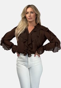 Brązowa bluzka z falbanami, z przezroczystymi rękawami, dekoltem w serek i zapięciem na guziki, zestawiona z białymi dżinsami o wysokim stanie. Gładka faktura materiału.