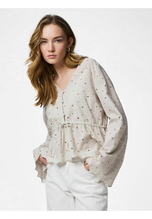 Tânără cu păr lung poartă o bluză bej deschis cu ochiuri și talie cu șnur, și pantaloni albi, pozând cu mâinile în buzunare.