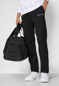 Schwarze Jogginghosen mit Seitentaschen und einem weißen Logo. Eine schwarze Sporttasche mit Mesh-Details wird in einer Hand gehalten. Weiße Sneakers runden den Look ab.
