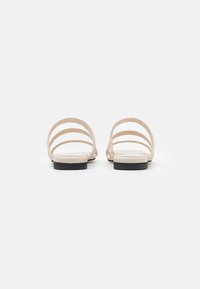 Sandali piatti beige con tre sottili cinghie curve sulla parte superiore, completati da una suola in gomma nera e una superficie liscia.