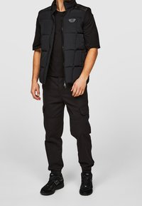 Schwarze Steppweste mit hohem Kragen und Frontreißverschluss, kombiniert mit einem schwarzen T-Shirt und Cargohosen. Verfügt über gepolsterte Bereiche und Logodetails.