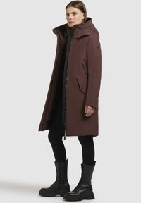 Brun vandtæt parka med høj krave, lynlås foran og sidelommer. Har et elegant design og er parret med sorte kampstøvler.