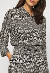 Chemise boutonnée avec un motif ondulé noir et crème ; manches longues, col, et taille élastique avec un détail noué. Tissu texturé.