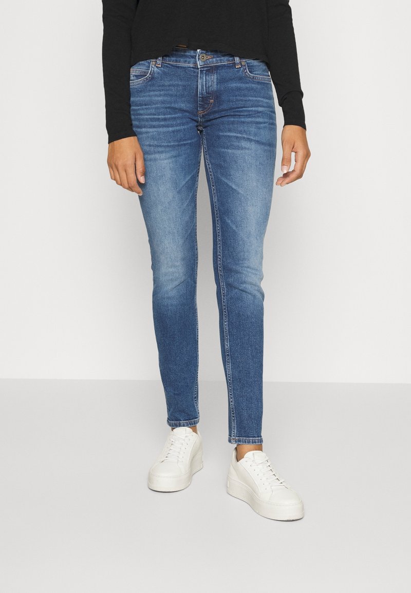 Person i blå slim-fit jeans, vita plattformssneakers och en svart långärmad topp, stående mot en ljus bakgrund.