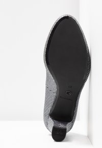 Semelle d'une chaussure à motifs reposant contre un mur d'angle blanc, montrant un dessous lisse noir avec la taille 37 et un talon à semelle antidérapante.
