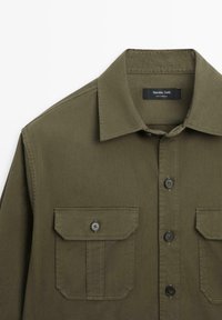 Camicia verde oliva con bottoni, colletto appuntito e due tasche sul petto con bottoni, vista della parte superiore anteriore e etichetta all'interno del colletto.