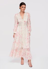 Femme debout portant une longue robe blanche à imprimé floral rose, bordure en dentelle, manches bouffantes et talons nus à brides, sur fond uni.