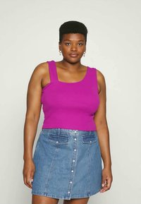 Mulher com cabelo curto, vestindo uma blusa sem mangas de um roxo brilhante e uma saia de ganga azul com botões, em pé contra um fundo claro e simples.