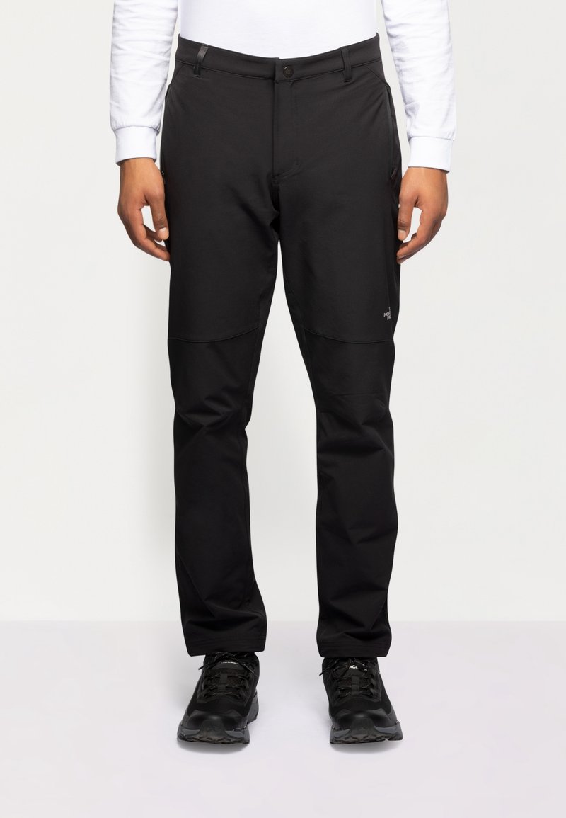 Homme portant un pantalon d'extérieur noir et des chaussures de randonnée noires, debout devant un fond blanc uni.