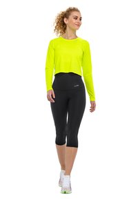 Neon-gelbes, kurzärmeliges Oberteil mit langen Ärmeln, kombiniert mit hochtaillierten schwarzen Capri-Leggings. Die Schuhe sind weiß mit bunten Akzenten.