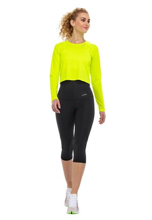 Neon-gelbes, kurzärmeliges Oberteil mit langen Ärmeln, kombiniert mit hochtaillierten schwarzen Capri-Leggings. Die Schuhe sind weiß mit bunten Akzenten.