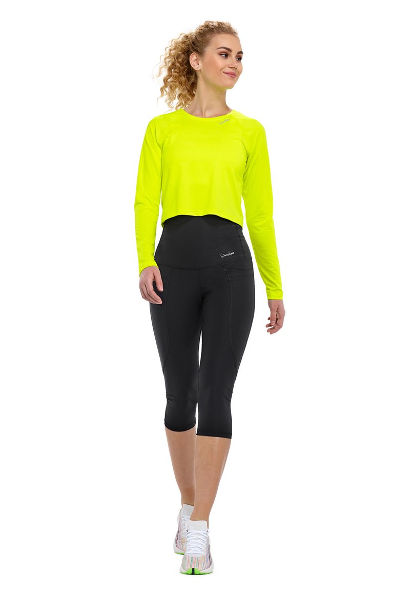 Neon-gelbes, kurzärmeliges Oberteil mit langen Ärmeln, kombiniert mit hochtaillierten schwarzen Capri-Leggings. Die Schuhe sind weiß mit bunten Akzenten.