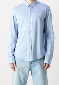 Mann trägt hellblaues, langärmliges Hemd mit Knöpfen und "BOSS"-Logo auf der Brust sowie hellblaue Jeans, steht vor weißem Hintergrund.