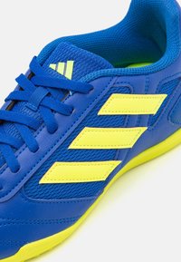 Blå och gula sportskor med blå snören, gul sula och tre gula ränder på sidan, vinklade på en vit bakgrund.