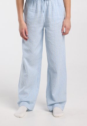 Persoon die losse, lichtblauwe en witte verticaal gestreepte broek draagt, blootsvoets staand tegen een effen achtergrond.