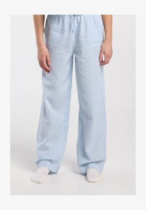 Persoon die losse, lichtblauwe en witte verticaal gestreepte broek draagt, blootsvoets staand tegen een effen achtergrond.