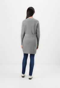 Szary długi kardigan z paskiem w talii, pleciony, z okrągłym dekoltem. Z tyłu znajduje się ozdobny guzik. Noszony na niebieskie dżinsy.