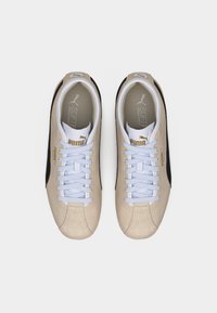 Μπεζ και μαύρα αθλητικά παπούτσια Puma με λευκά κορδόνια, χρυσό λογότυπο στο πλάι και στη γλώσσα, εμφανιζόμενα από πάνω σε λευκό φόντο.