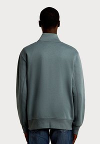 Szaro-niebieska bluza z półgolfem, luźnym krojem i ściągaczami na rękawach. Charakteryzuje się widocznym przeszyciem i gładką fakturą.