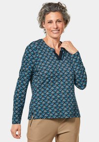Langärmliges Oberteil in Blau mit einem geometrischen Muster aus Kreisen. V-Ausschnitt-Design mit kontrastierenden marineblauen Akzenten und seitlichen Schlitzen für mehr Komfort.
