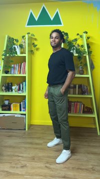 Musta lyhythihainen t-paita, vihreät cargo-housut ja valkoiset lenkkarit. Keltainen seinä, jossa vihreä vuorimainen kuvio, ja vihreitä hyllyjä, joissa kirjoja ja kasveja.