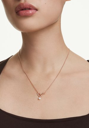 Zbliżenie szyi kobiety, na której wisi delikatny naszyjnik z różowego złota z zapięciem w kształcie serca oraz małym białym perłowym zawieszeniem.