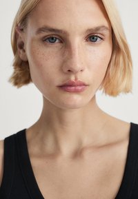 Portret osoby z jasnym blond bobem, wyraźnymi niebieskimi oczami, piegami i gładką skórą, ubraną w czarną bezrękawnik.