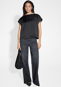 Schwarzes Satinoberteil mit kurzen Ärmeln und weitem Ausschnitt; kombiniert mit dunklen Denim-Jeans im weiten Schnitt. Das Model hält eine schwarze strukturierte Tasche und trägt spitze Absätze.