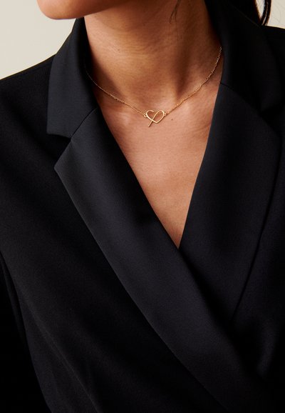 Collana d'oro con ciondolo a forma di cuore, indossata sopra un top di tessuto nero con scollatura a V. Catena fine con una texture liscia.