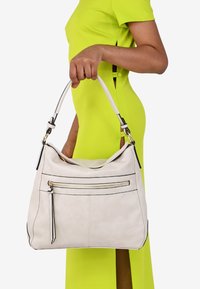 Borse a tracolla in pelle color beige chiaro con dettagli neri, tasca con zip e tracolla curva, appoggiata contro un vivace vestito giallo.