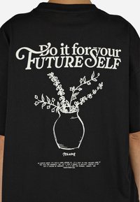 Czarna koszulka z krótkim rękawem z białym napisem i prostą ilustracją wazonu z liśćmi i kwiatami, głównie umieszczoną na plecach.