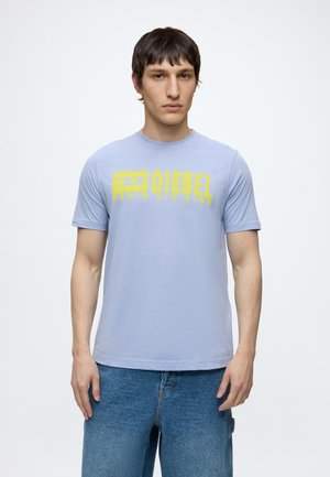 Jeune homme portant un t-shirt bleu clair avec le logo jaune "DIESEL" et un jean bleu, debout devant un fond blanc uni.
