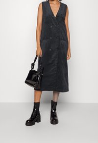 Musta, v-aukkoinen midimittainen corduroy-mekko, jossa on kaksiriviset napit ja sivutaskut, yhdistetty mustaan teksturoituun käsilaukkuun ja paksupohjaisiin saappaisiin.