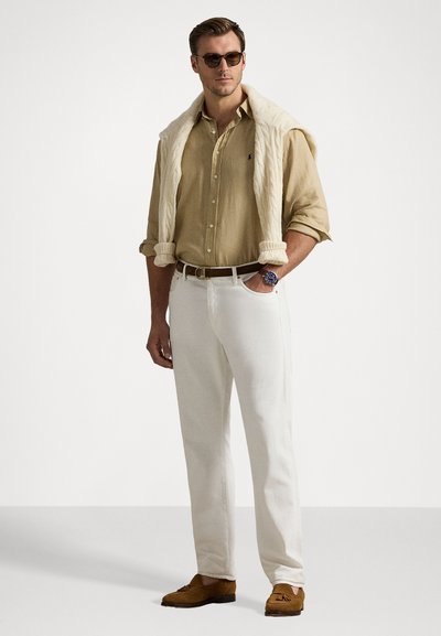 Camisa bege de linho com botões, calças brancas, suéter de malha creme colocado sobre os ombros, sapatos moccasins castanhos e óculos de sol.