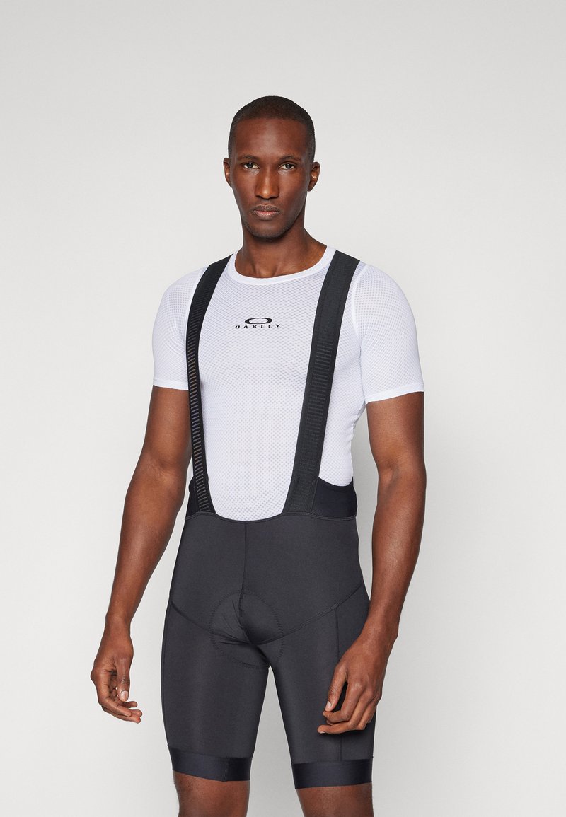 Juodos dviračių šortai su reguliuojamais petnešomis, vilkint baltą tinklelio marškinėlį. Su paminkštinta sėdimosios dalies zona ir logotipu marškinėlių priekyje.