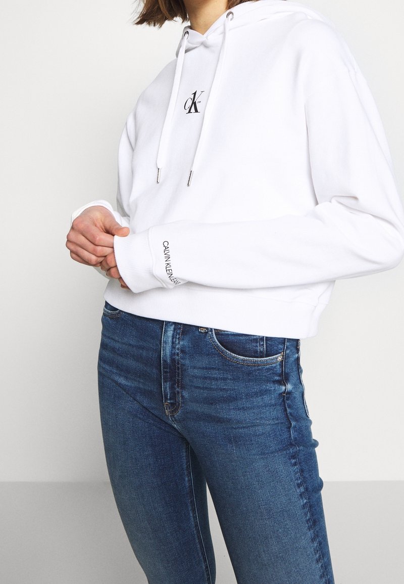 Witte cropped hoodie met een capuchon en trekkoord en een zwart logo op de borst. Gecombineerd met nauwsluitende blauwe spijkerbroek met een klassiek ontwerp.