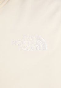 Beige látka se vyšitým logem "The North Face" v bílé barvě, s křivkovým designovým prvkem nad textem.