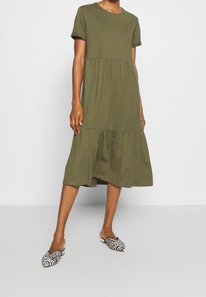 Nainen pukeutuneena oliivinvihreään lyhythihaiseen midimekkoon, jossa on röyhelöhelma, yhdistetty mustavalkoisiin kuvioituihin slip-on-kenkiin.