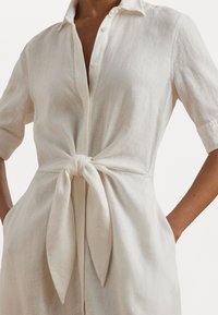 Light beige linen shirt dress with a collar, short sleeves, and a tied waist. Features a buttoned front and soft, textured fabric.