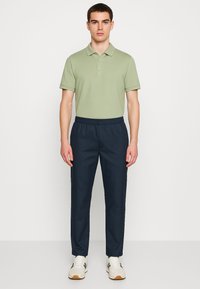 Polo shirt verde chiaro a maniche corte, abbinato a pantaloni navy con vita elastica. Le scarpe sono bianche con dettagli verdi e suole marroni.