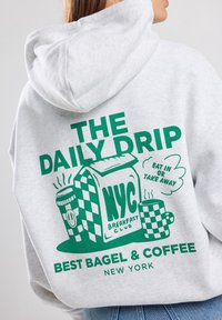 Grå huvtröja med en stor grön grafik på baksidan, som föreställer kaffe och bagel, med texten "THE DAILY DRIP" och "BEST BAGEL & COFFEE."