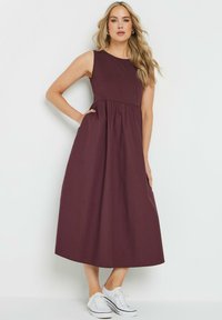 Vestido sin mangas en color burdeos con un busto ajustado y falda fruncida. Diseñado en tejido suave, combinado con zapatillas blancas para crear contraste.