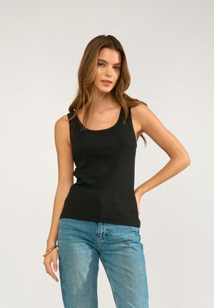 Giovane donna con top nero senza maniche e jeans blu in piedi davanti a uno sfondo semplice con una mano sul fianco.
