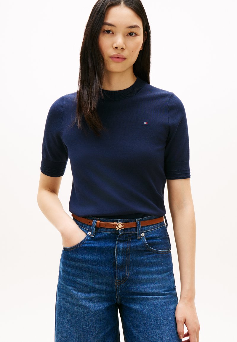 Jeune femme portant un haut bleu marine à manches courtes, une ceinture marron et un jean taille haute bleu, debout avec une main dans la poche.