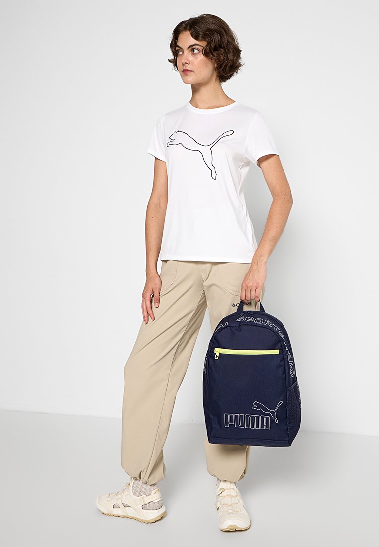 Camiseta blanca con un logo negro de Puma, pantalones beiges y una mochila azul marino con acentos en amarillo neón y el logo de Puma.