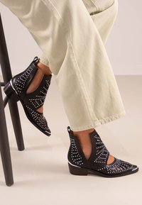 Botines de cuero negros con diseños troquelados y tachuelas plateadas. El tacón es bajo, con punta afilada y acentos brillantes.