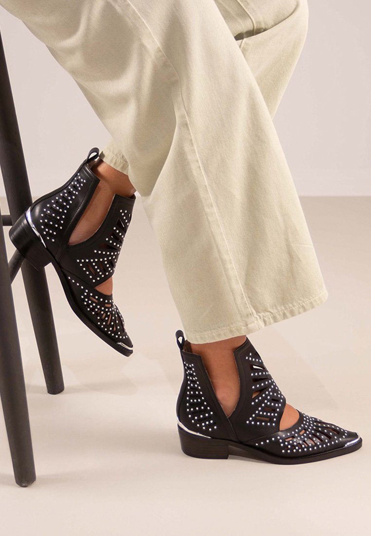 Botines de cuero negros con diseños troquelados y tachuelas plateadas. El tacón es bajo, con punta afilada y acentos brillantes.