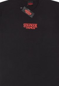 Černé bavlněné tričko s červeným logem "Stranger Things" ve zrezivělém písmu na přední straně. Design s kulatým výstřihem. S připevněnou etiketou.