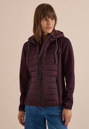 Kvinde iført en vinrød quiltet hættejakke med frontlynlås og blå jeans, stående foran en ensfarvet beige baggrund.