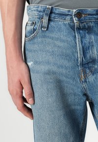 Jeans de mezclilla azul claro con un acabado desteñido, diseño de cinco bolsillos, costuras naranjas y un cierre de botón en la cintura.